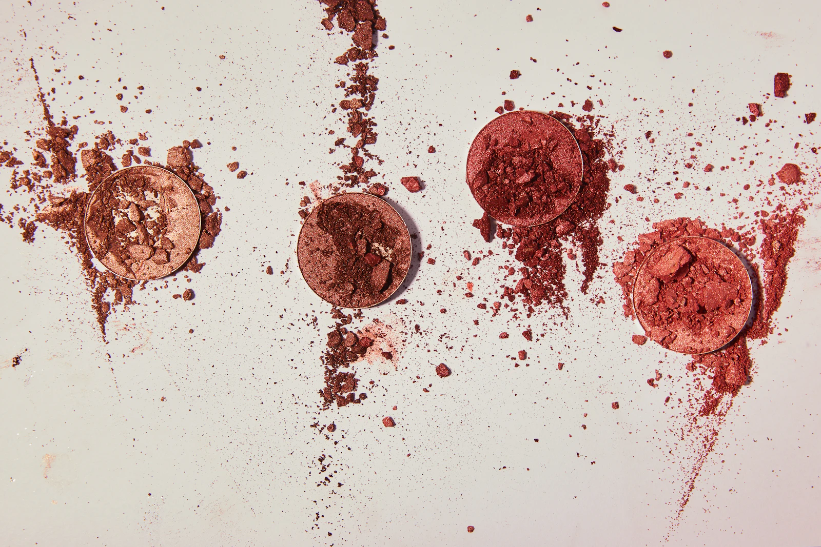 Crushed pressed powders in red, rose, and copper tones scattered across a pale surface