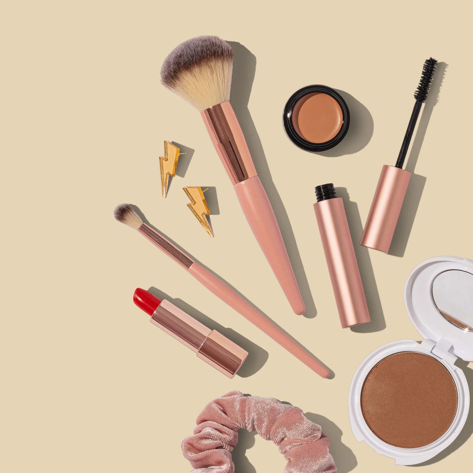Flat lay of two brushes, mascara, a red lipstick, and bronzer on a sand-colored background