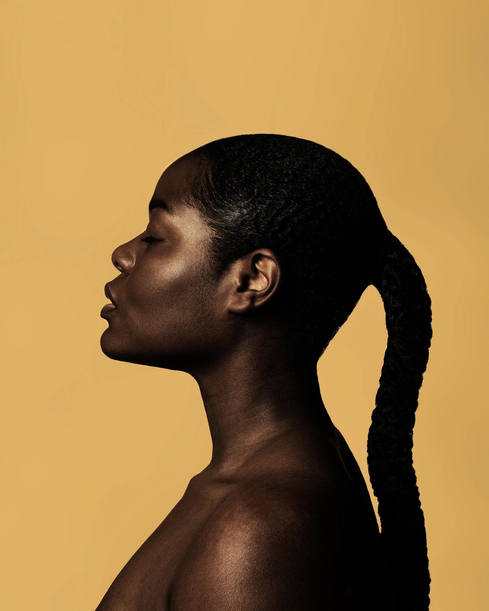 Profile of a woman with deep skin and a long braided ponytail against an ochre wall