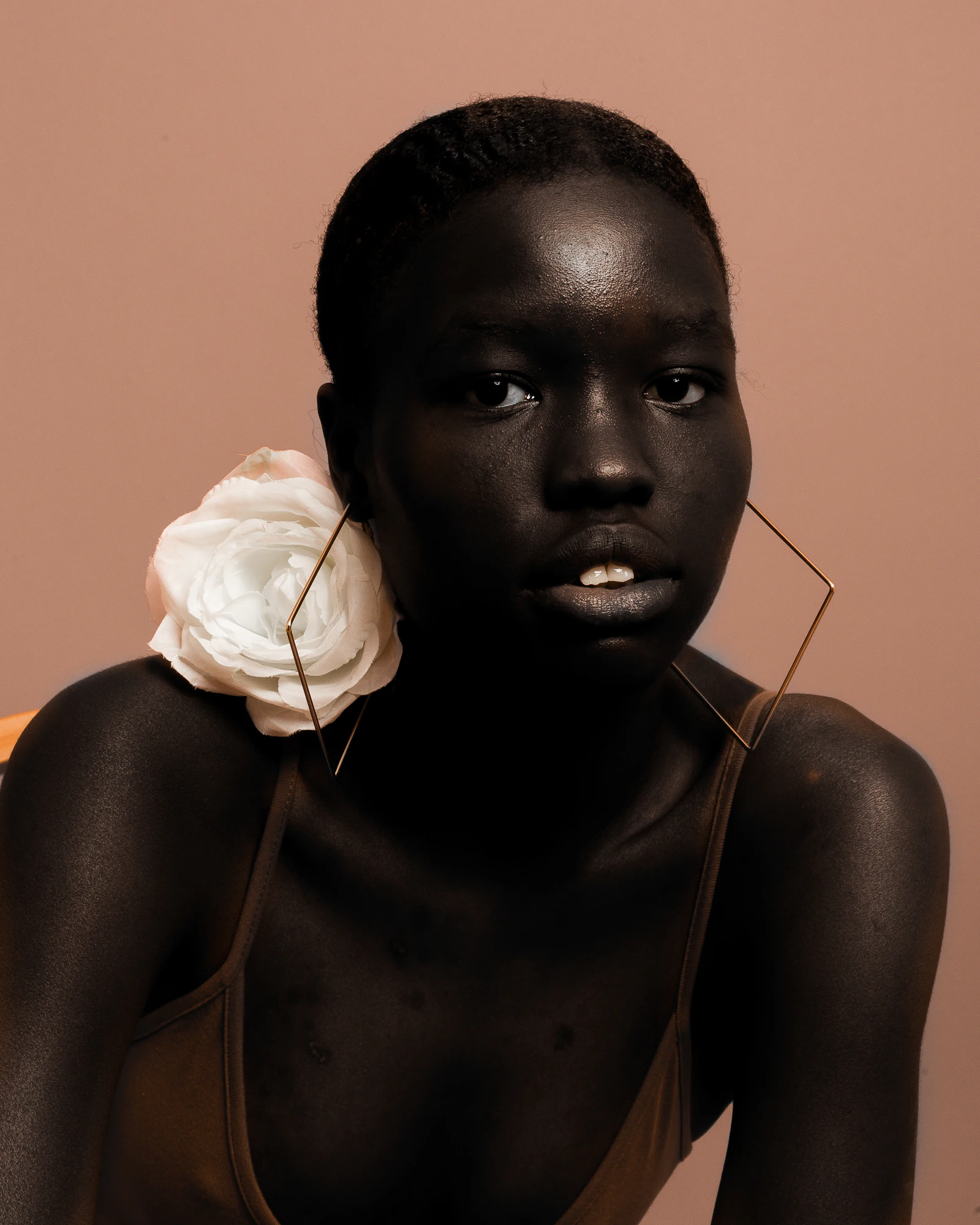 Woman with deep skin wearing a white rose behind her ear against a blush-colored wall