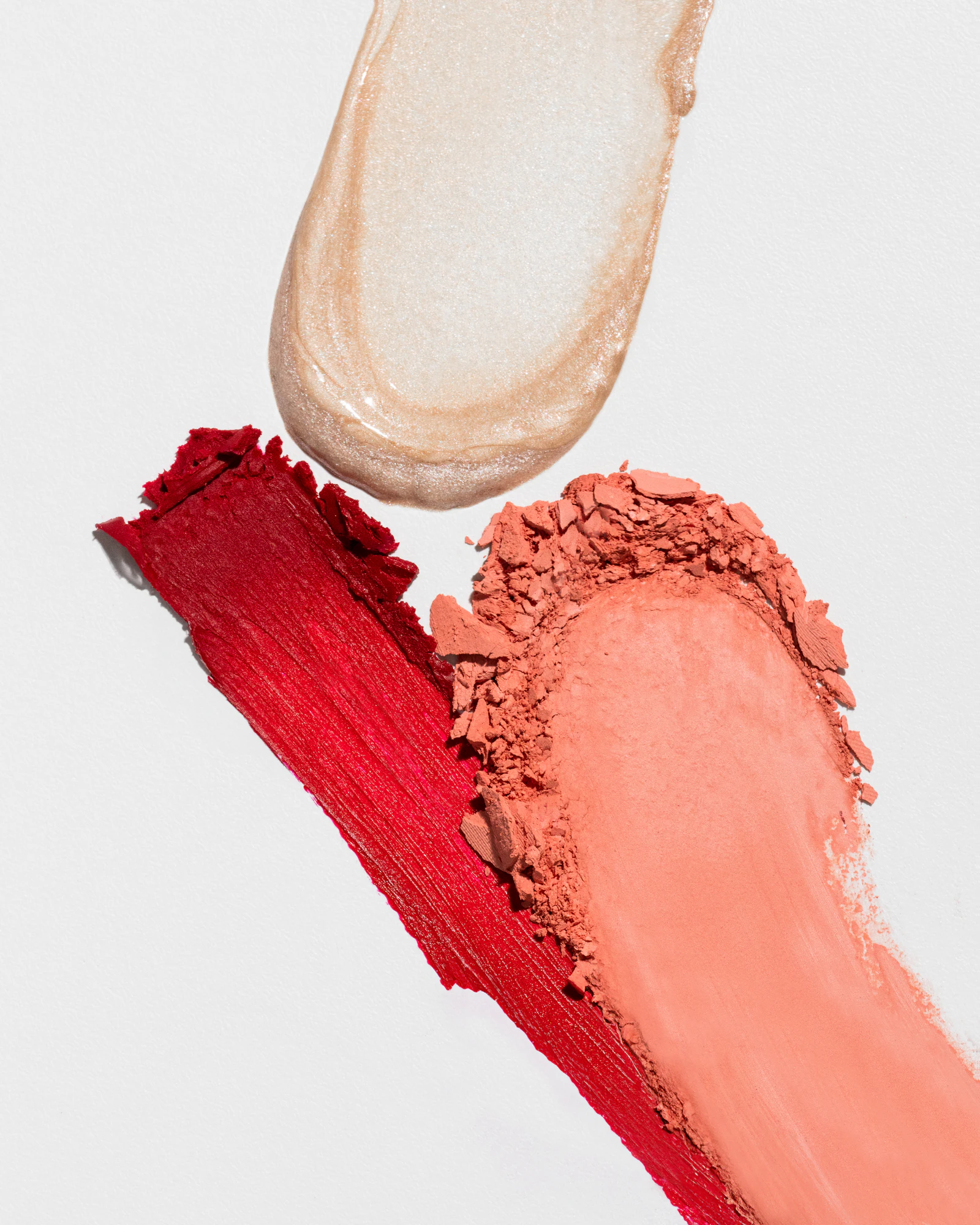 Smears of shimmer highlighter, red lip color, and crumbled peach blush on white